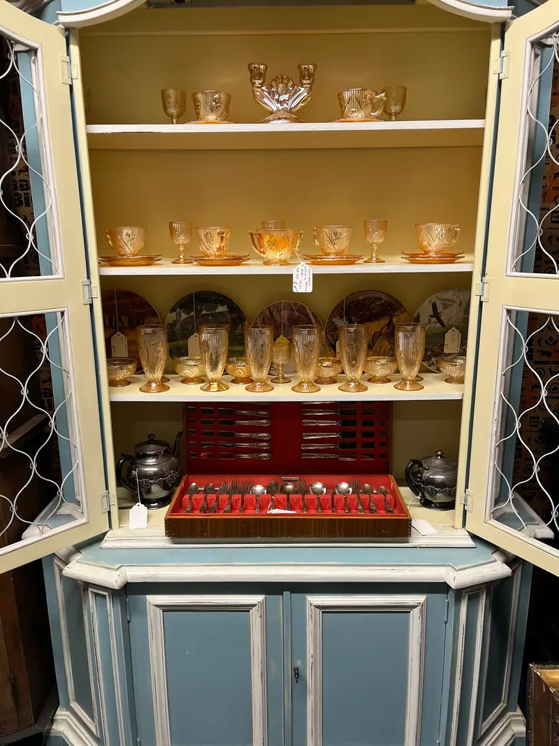 A blue-painted china cabinet filled with amber glassware and a silverware chest